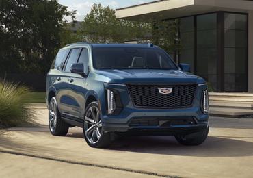 Front ¾ view of the 2025 Cadillac Escalade Sport Platinum in Deep Sea Metallic with a house and greenery in the background.